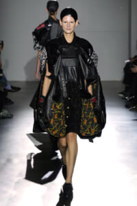 Model walks the runway in an avant-garde black ensemble: oversized leather cape, textured black top, and a skirt with a sheer, dark floral panel; glossy headpiece completes the look.