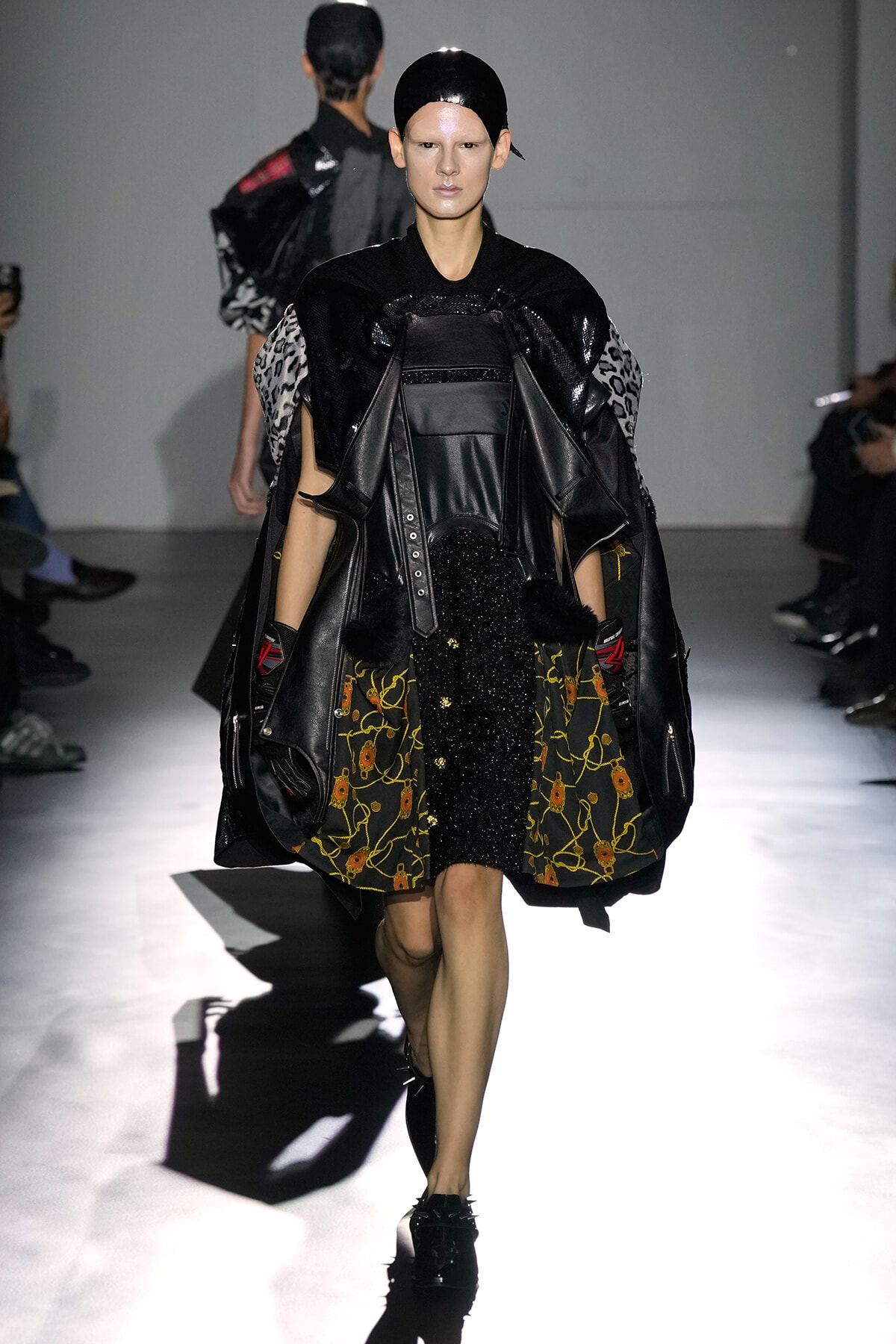 Model walks the runway in an avant-garde black ensemble: oversized leather cape, textured black top, and a skirt with a sheer, dark floral panel; glossy headpiece completes the look.