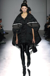 Model walking a runway in an oversized black utility cape with reflective band details and glossy leggings, holding a beaded bag.