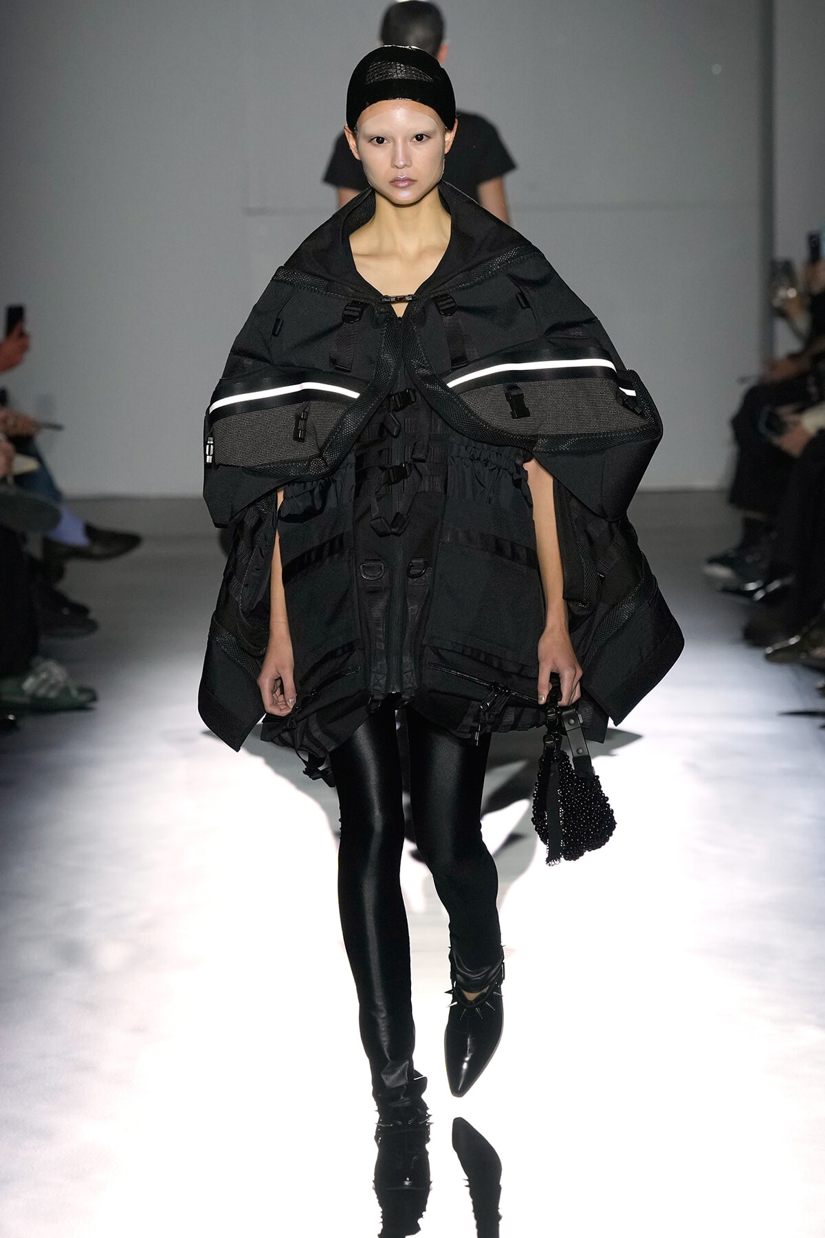 Model walking a runway in an oversized black utility cape with reflective band details and glossy leggings, holding a beaded bag.