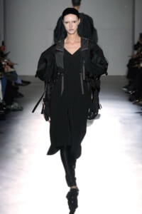 Model walking runway in a black, oversized, utilitarian dress with large puffed sleeves and harness-like straps.