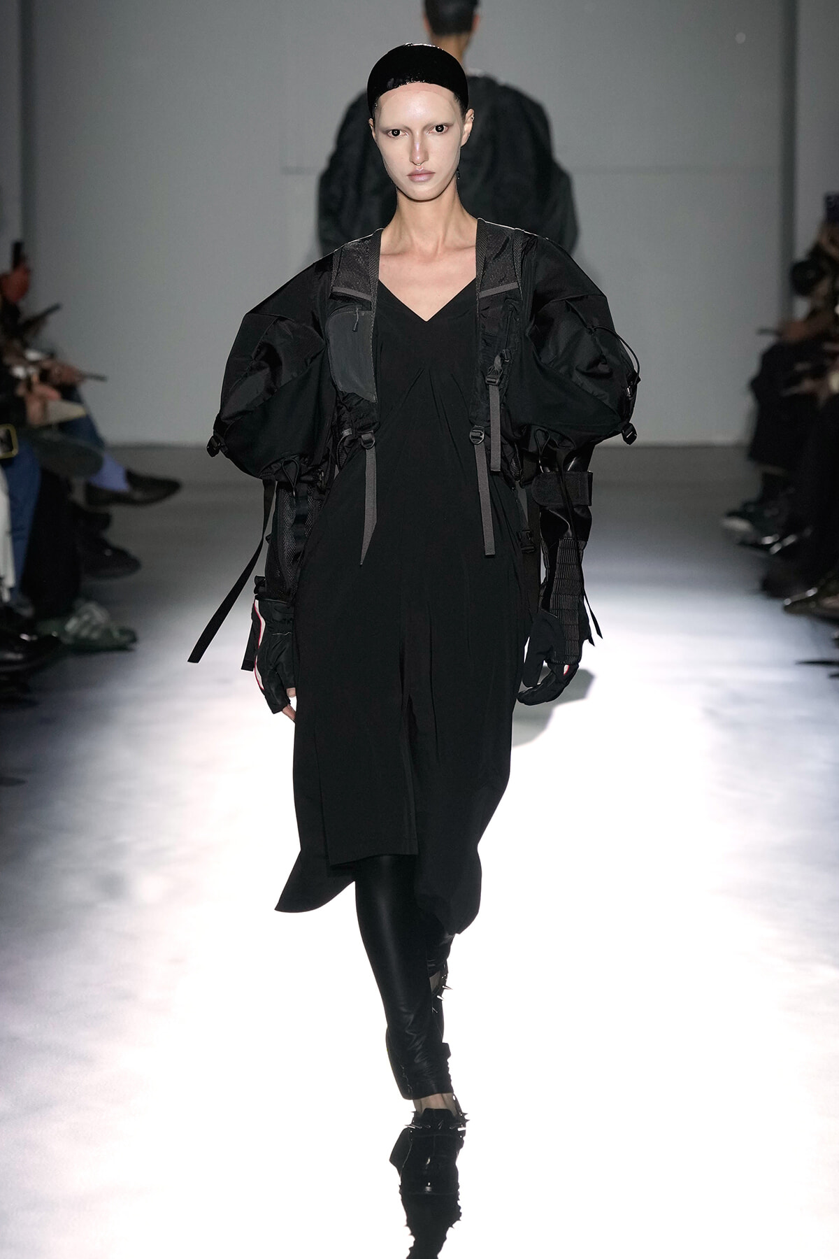 Model walking runway in a black, oversized, utilitarian dress with large puffed sleeves and harness-like straps.