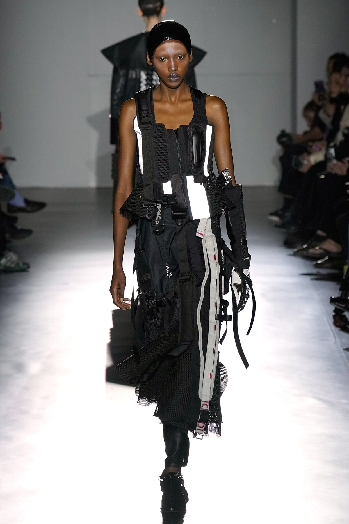 Female model on a runway wearing an industrial, strap-filled black-and-white outfit with buckles and utility pockets.