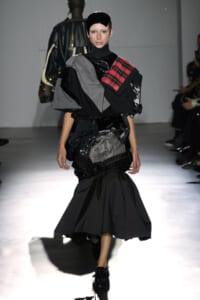 Model on a runway wearing a dramatic black layered outfit with gray and plaid shoulder accents and a black cap, mid-stride.