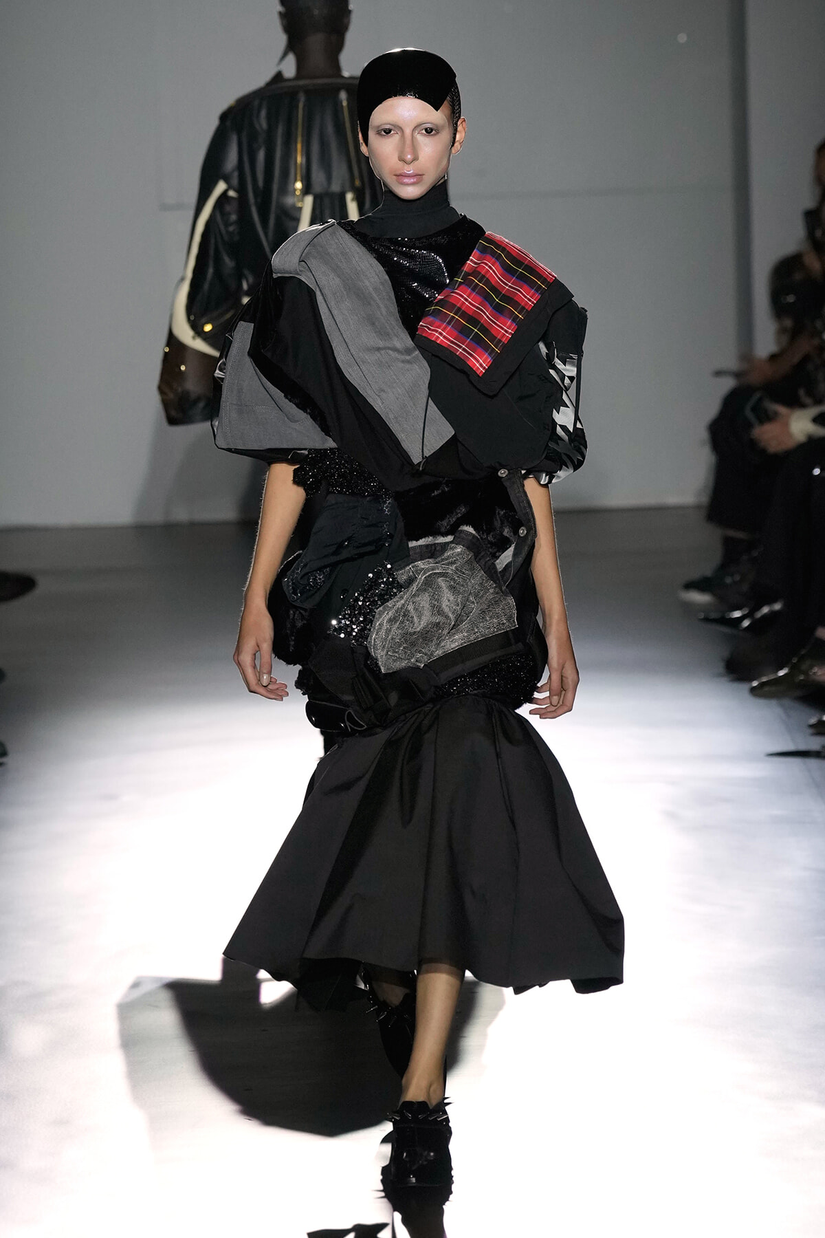 Model on a runway wearing a dramatic black layered outfit with gray and plaid shoulder accents and a black cap, mid-stride.