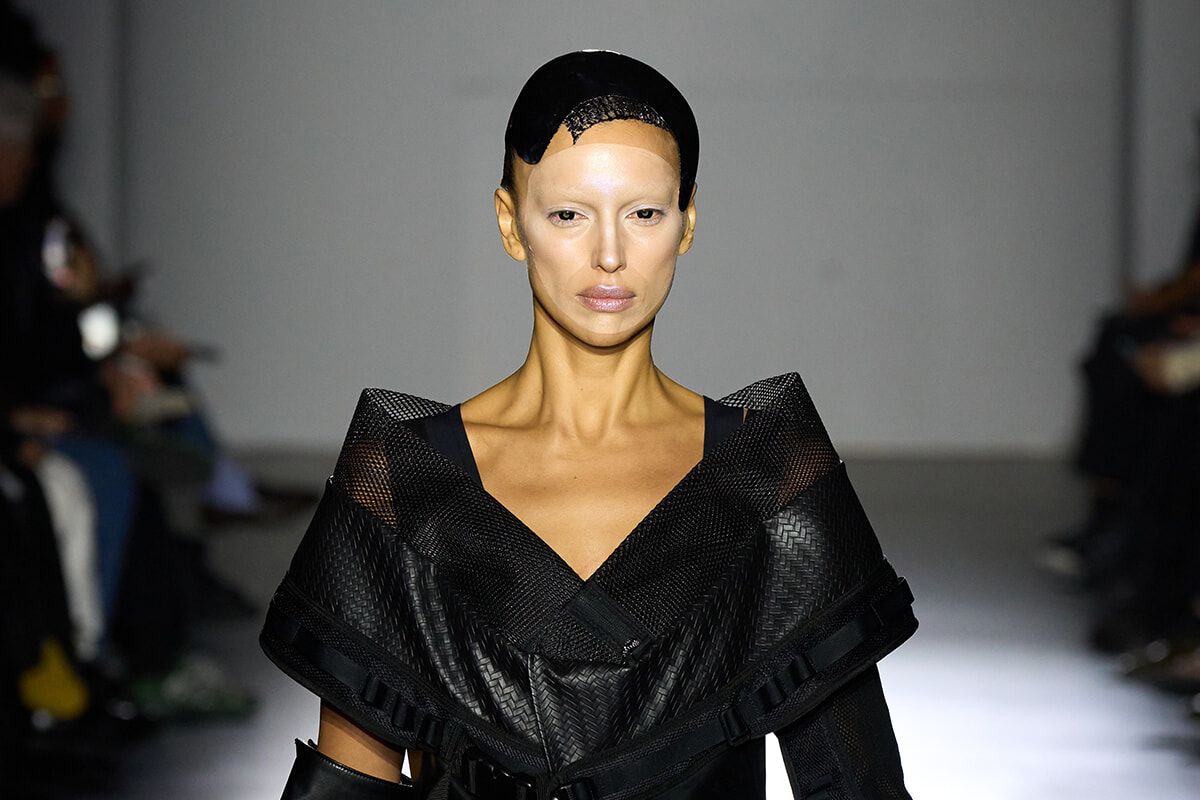 Model walks the runway in a dramatic black geometric cape with oversized angular shoulders, under dim studio lighting.