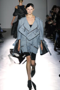 Model walks the runway in a voluminous blue-gray trench with multiple zippers and pockets on display.