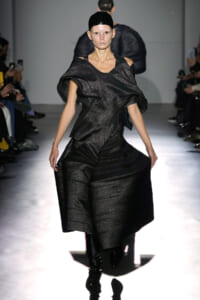 Model walks the runway in a black metallic off-the-shoulder sculptural gown with voluminous sleeves and a fitted waist, spotlighted on a glossy catwalk.