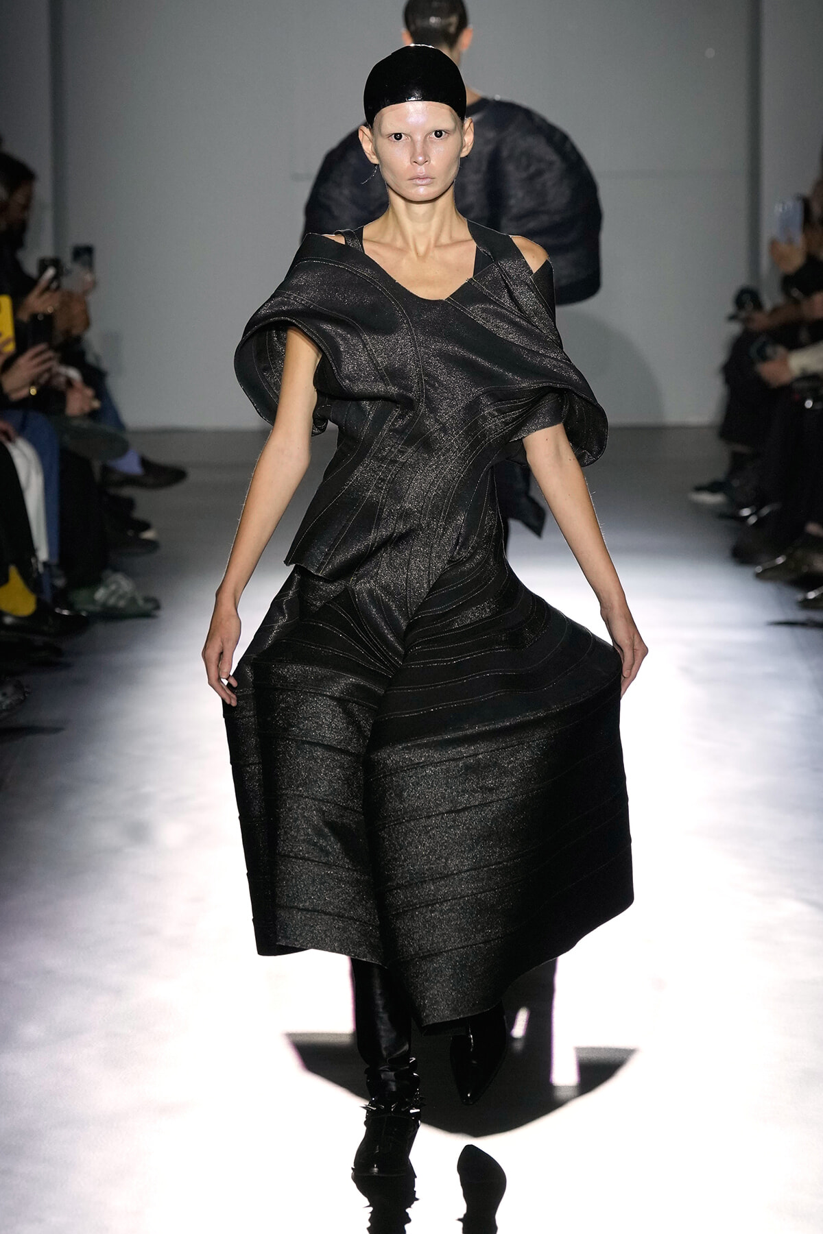 Model walks the runway in a black metallic off-the-shoulder sculptural gown with voluminous sleeves and a fitted waist, spotlighted on a glossy catwalk.