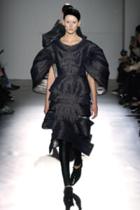 Model walks the runway in a dark metallic, sculpted dress with oversized sleeves and layered skirt.
