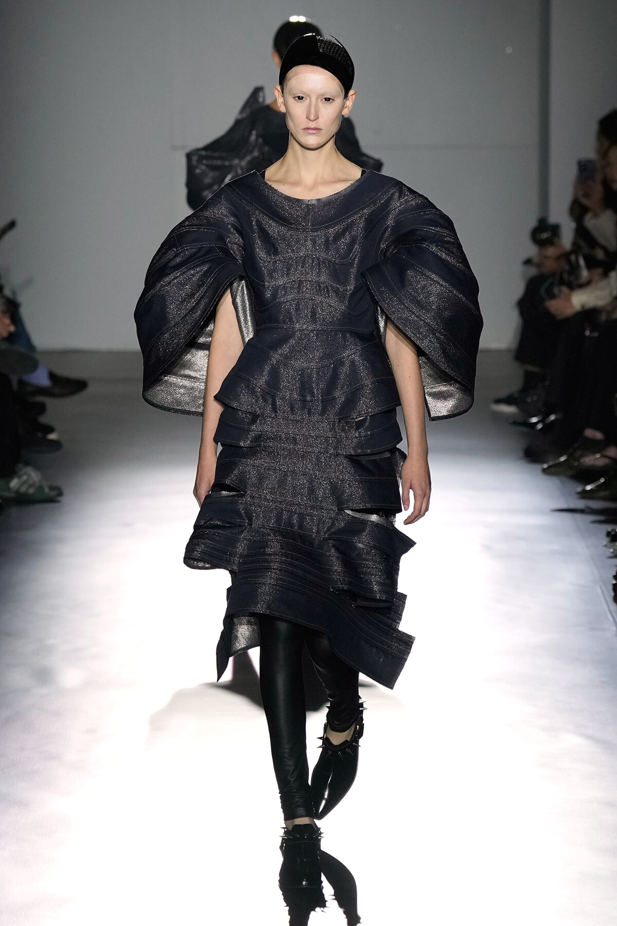 Model walks the runway in a dark metallic, sculpted dress with oversized sleeves and layered skirt.