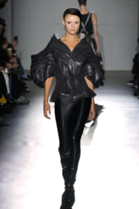 Model walks the runway in a sculptural black metallic top with huge puff sleeves and glossy black pants.