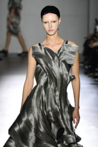 Model walks a runway in a metallic sculptural silver gown with angular shoulder details and a black cap.