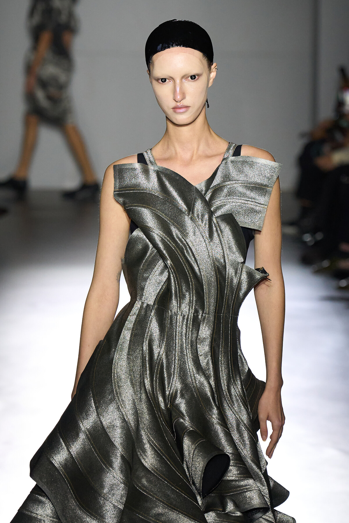 Model walks a runway in a metallic sculptural silver gown with angular shoulder details and a black cap.