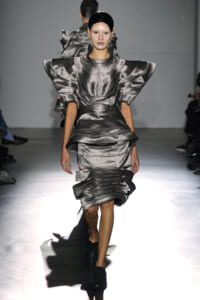 Model on a runway wearing a metallic silver sculptural dress with exaggerated angular shoulders and layered, ruffled tiers.