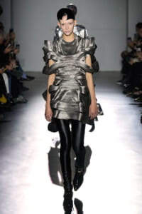 Model on a runway wearing a futuristic metallic silver dress with sculpted wrapped panels and black tights.