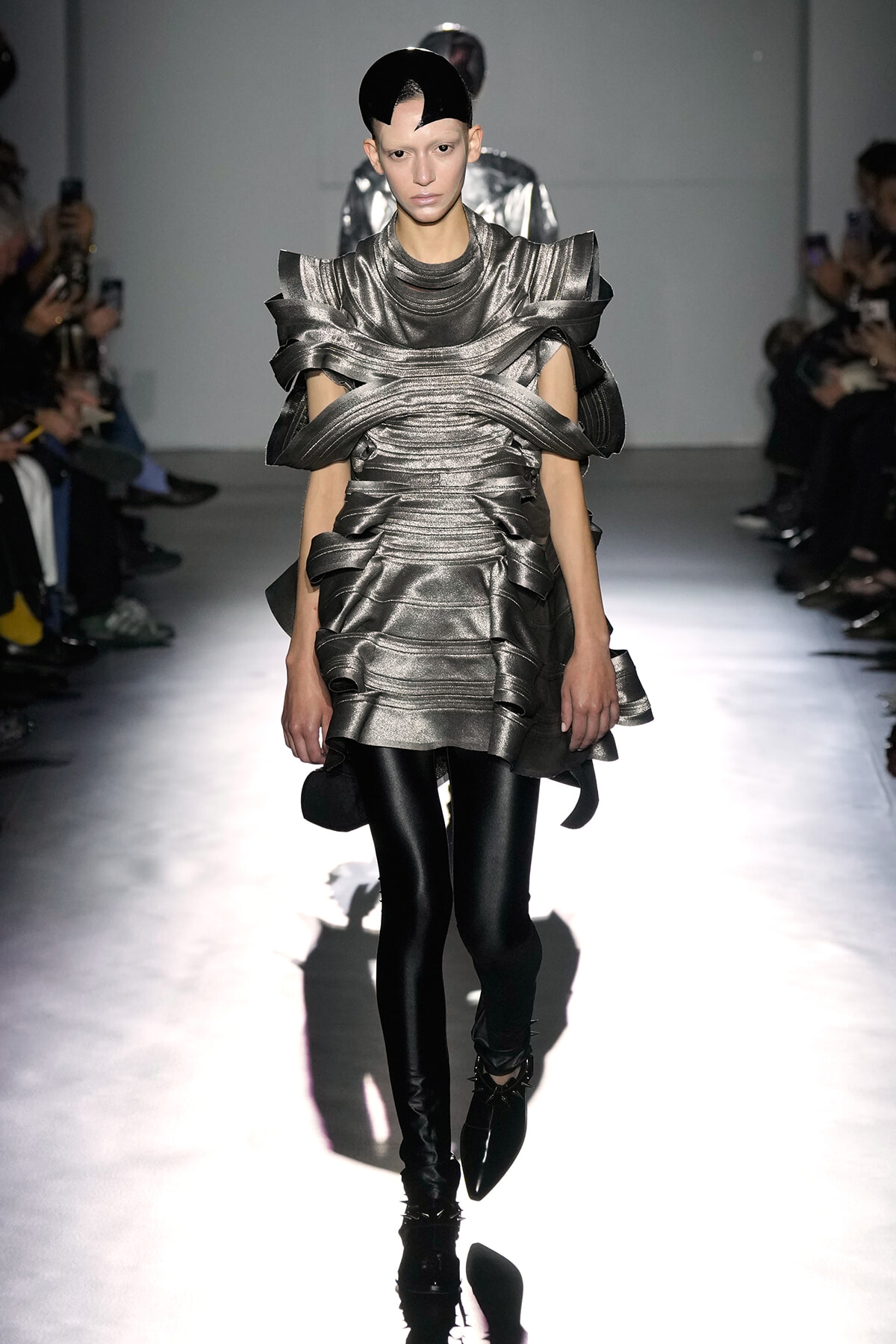 Model on a runway wearing a futuristic metallic silver dress with sculpted wrapped panels and black tights.