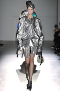 Model walking the runway in a metallic silver oversized jacket with matching shorts and a spiked, pearl-studded bag.