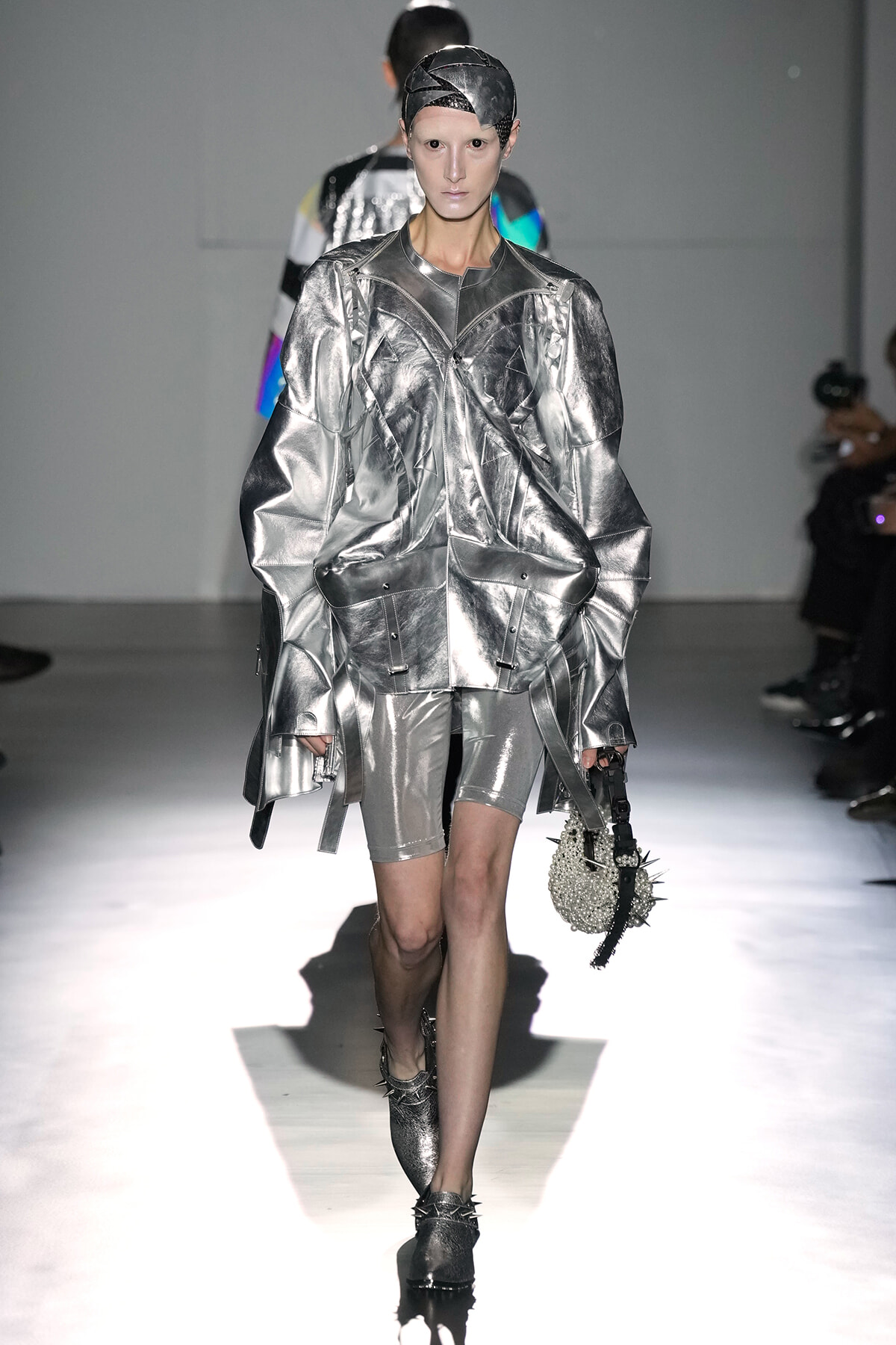 Model walking the runway in a metallic silver oversized jacket with matching shorts and a spiked, pearl-studded bag.