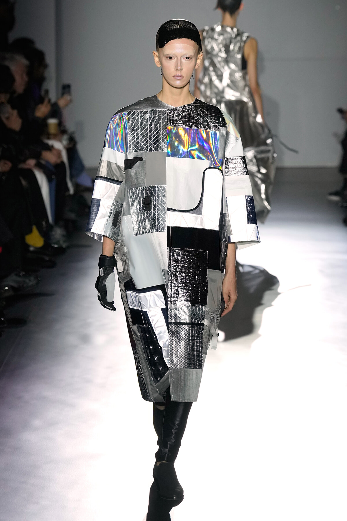 Model walks the runway in a futuristic patchwork metallic dress in silver, black, and holographic panels, with a black headband and gloves.