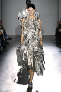 Model walks a metallic silver avant-garde outfit on a runway, with matching headgear and chunky boots amid a spotlighted catwalk.