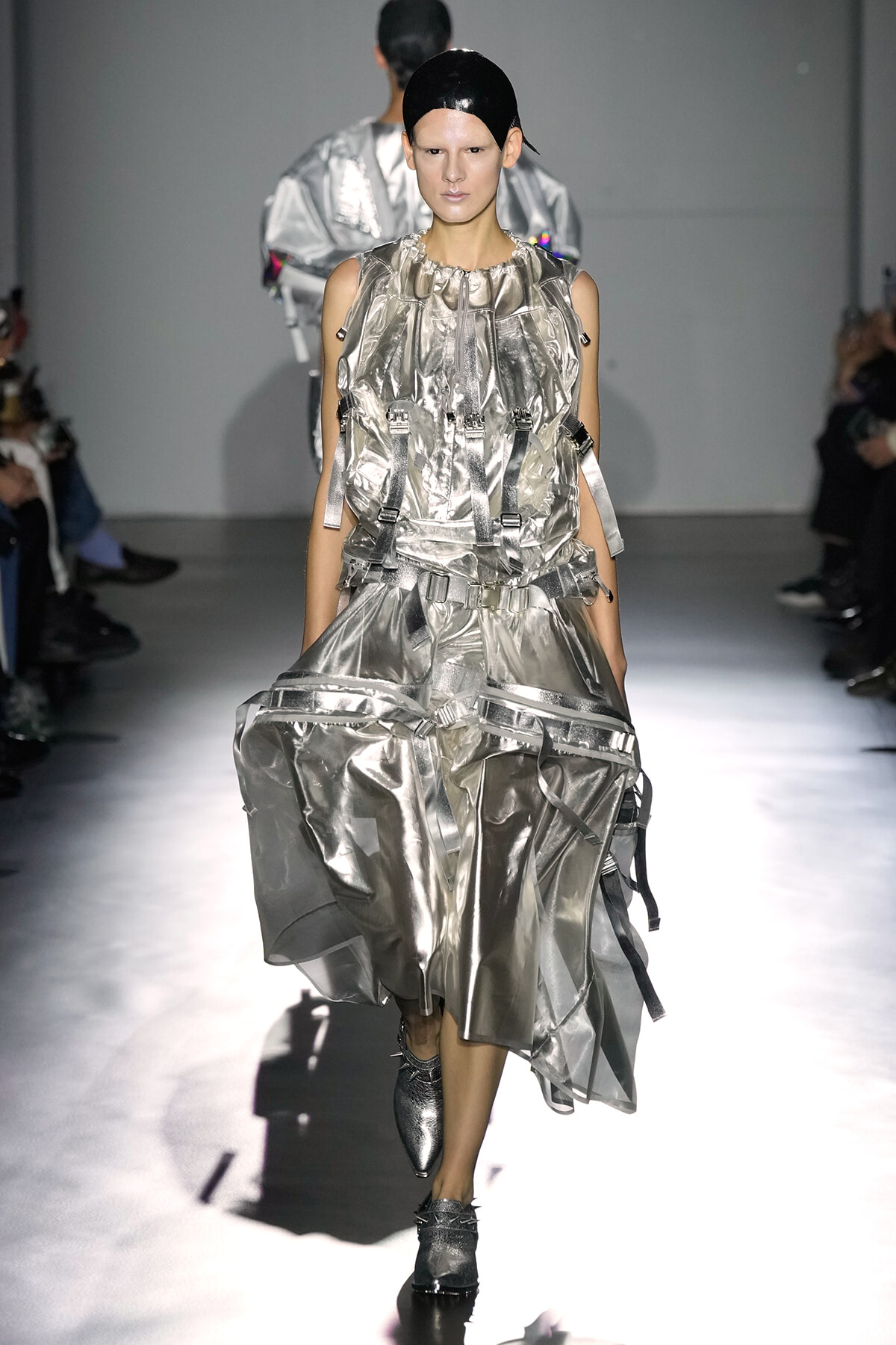 Model walks a metallic silver avant-garde outfit on a runway, with matching headgear and chunky boots amid a spotlighted catwalk.