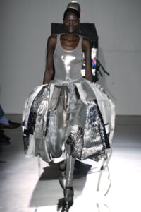 Model walking the runway in a silver metallic tank dress with a voluminous, patchwork skirt of reflective panels and straps, pairing with metallic boots.