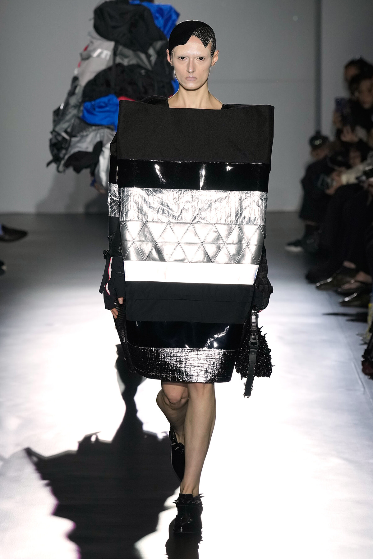 Model walks a fashion runway in a oversized black and silver geometric outfit with metallic panels; black headpiece with netting.