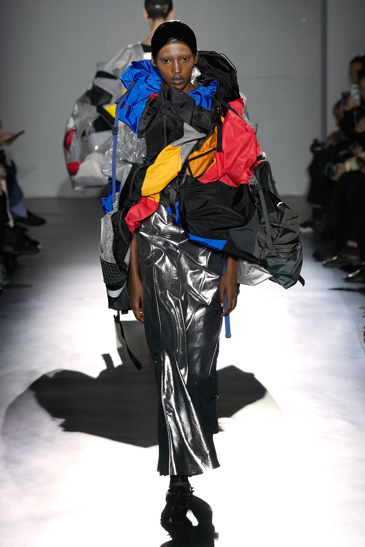 Runway model in a bold, oversized color-blocked outfit with a shiny silver metallic skirt, on a fashion show runway.