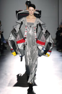 Model walks the runway in a metallic silver, oversized layered outfit with wing-like panels and colorful accents; futuristic fashion design.