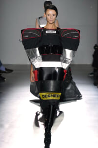 Model walks the runway in a futuristic black armor-inspired ensemble with silver panels, red piping, and a Degner label prominently displayed at the chest area.