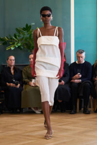 Model walks runway in white layered dress, pink sheer gloves, sunglasses, and statement earrings; audience seated in background.