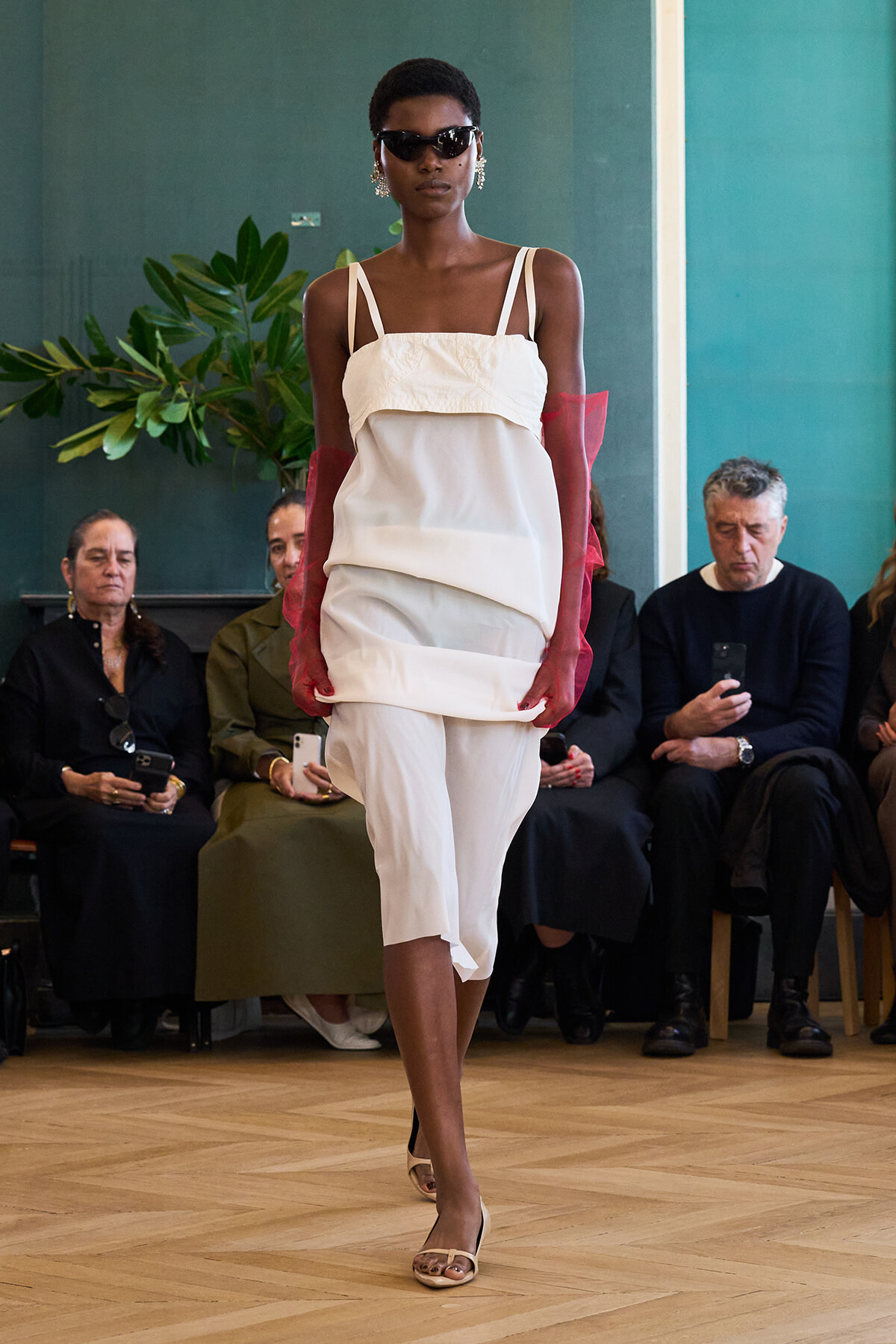 Model walks runway in white layered dress, pink sheer gloves, sunglasses, and statement earrings; audience seated in background.