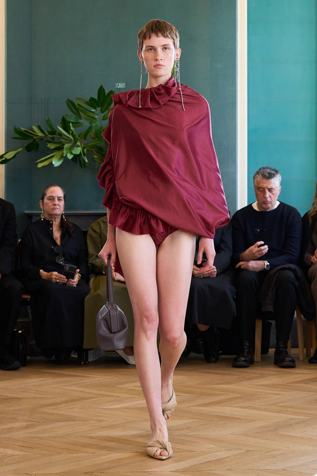 Model struts a burgundy draped one-shoulder top with ruffled hem and matching bottoms, carrying a small gray handbag; audience watches from seats in background.