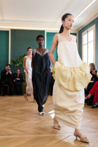 Model in a cream sleeveless dress with a large pale yellow draped skirt walks the runway, followed by two other models in contrasting gowns; audience seated beside a green wall.