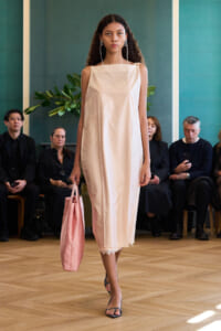 Model walks the runway in a pale pink sleeveless midi dress with lace hem, carrying a matching pink tote bag and wearing statement earrings in a fashion show setting.