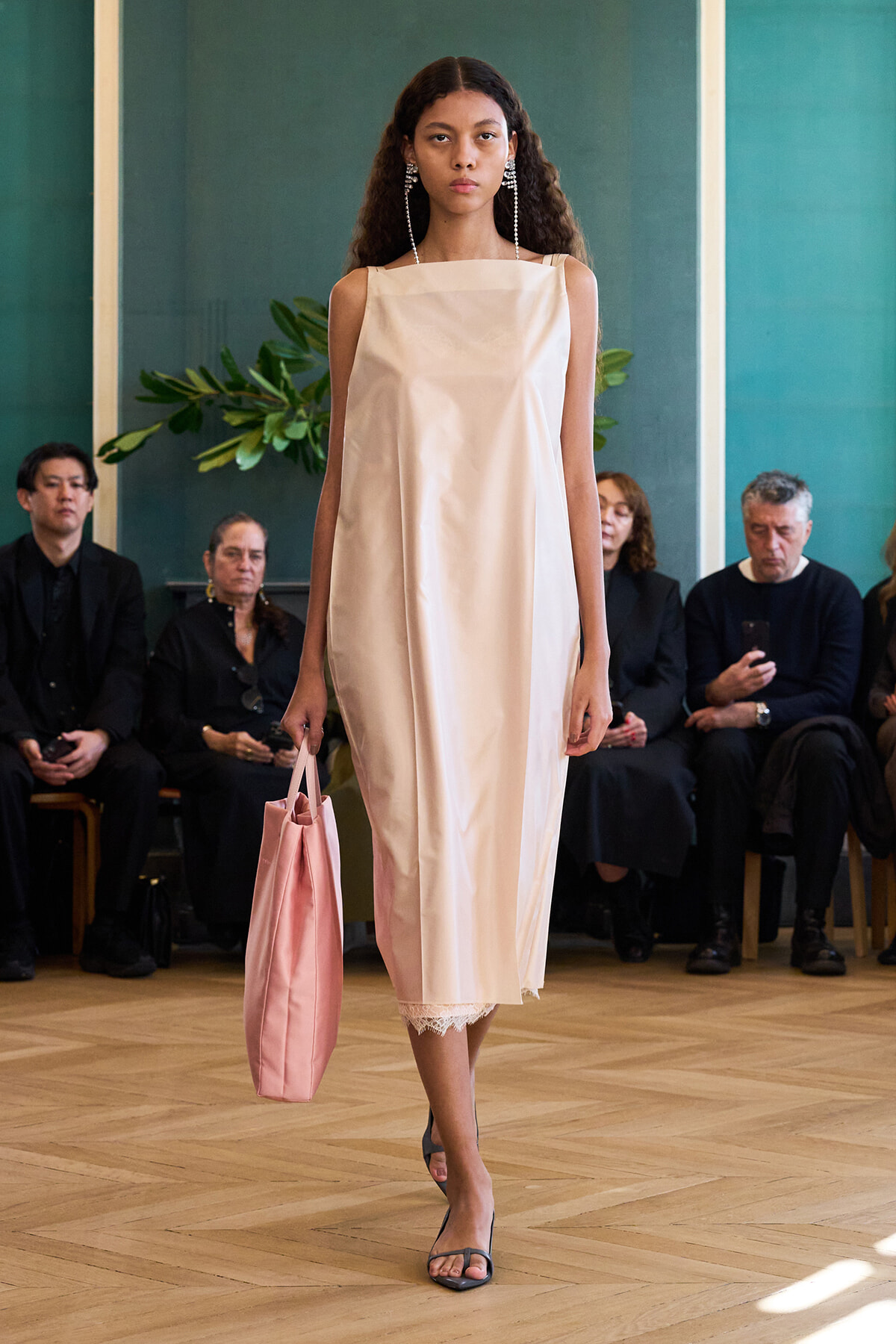 Model walks the runway in a pale pink sleeveless midi dress with lace hem, carrying a matching pink tote bag and wearing statement earrings in a fashion show setting.