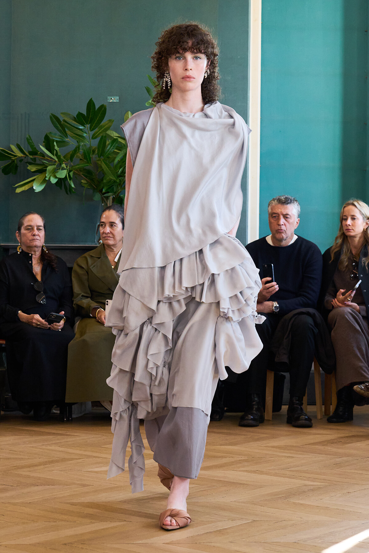 Model walks the runway in a light gray, layered draped dress with open-toed sandals; seated audience watches in a fashion show setting.