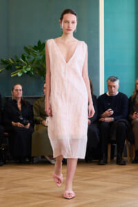 Model walks the runway in a pale pink sleeveless V-neck midi dress with a sheer hem; pink twisted-strap sandals and audience seated in the background.