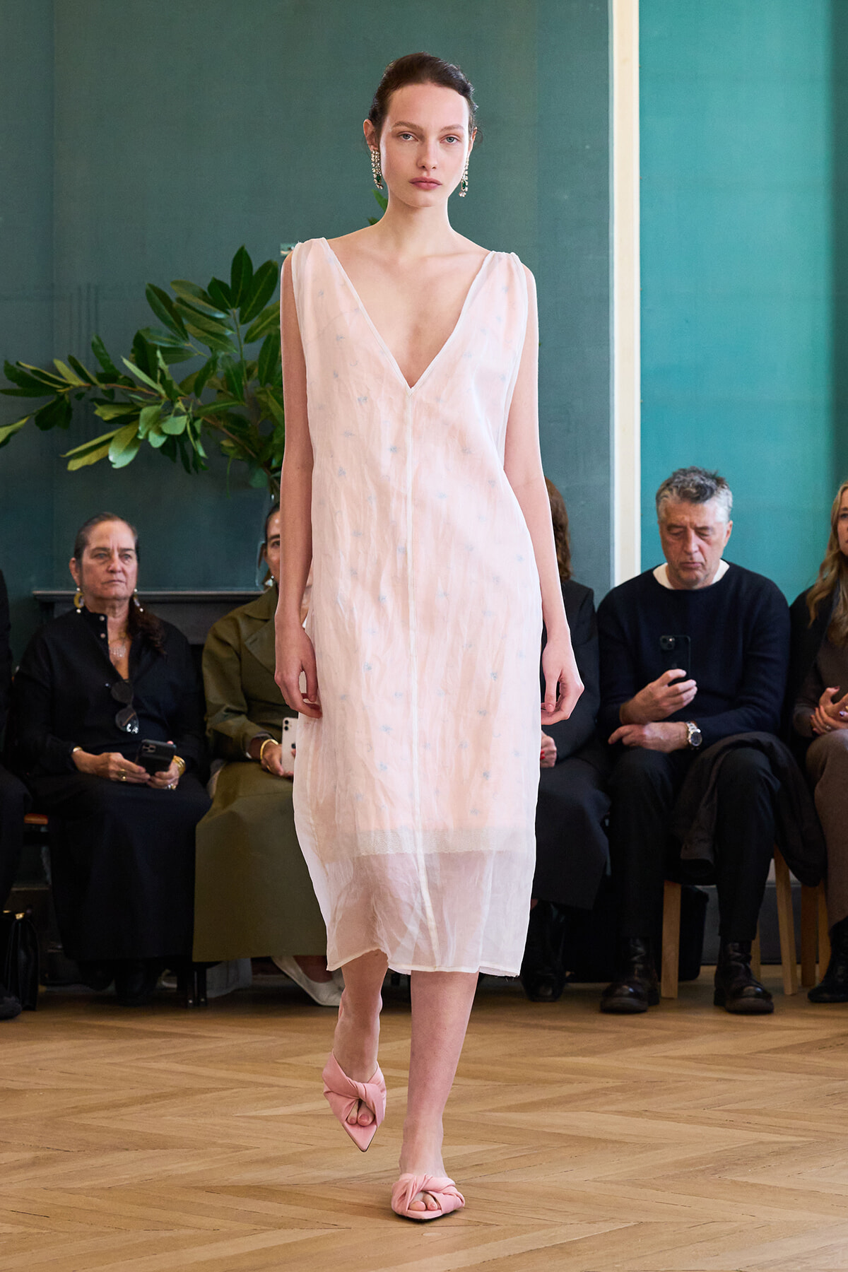 Model walks the runway in a pale pink sleeveless V-neck midi dress with a sheer hem; pink twisted-strap sandals and audience seated in the background.