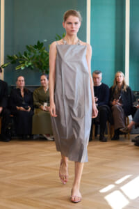 Model walks a silver-gray midi dress with a structured, square neckline and dangling necklace on a fashion show runway.