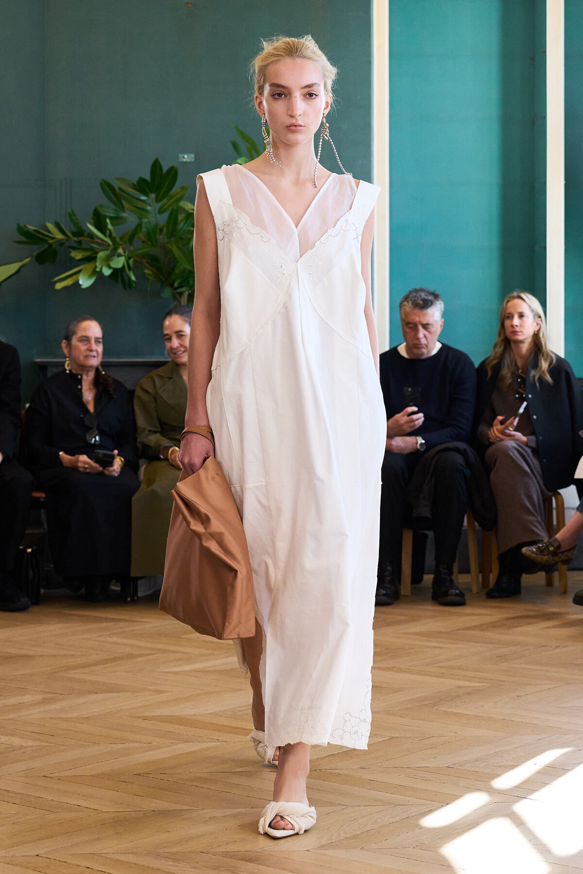 Model walks the runway in a white sleeveless maxi dress with delicate lace details, carrying a tan clutch as spectators watch from the audience.