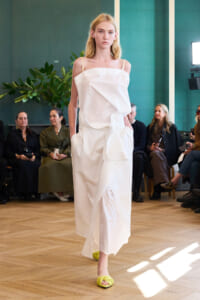 Model walks a fashion runway in a white draped dress, neon-yellow sandals, and a neutral expression, with seated audience in the background.