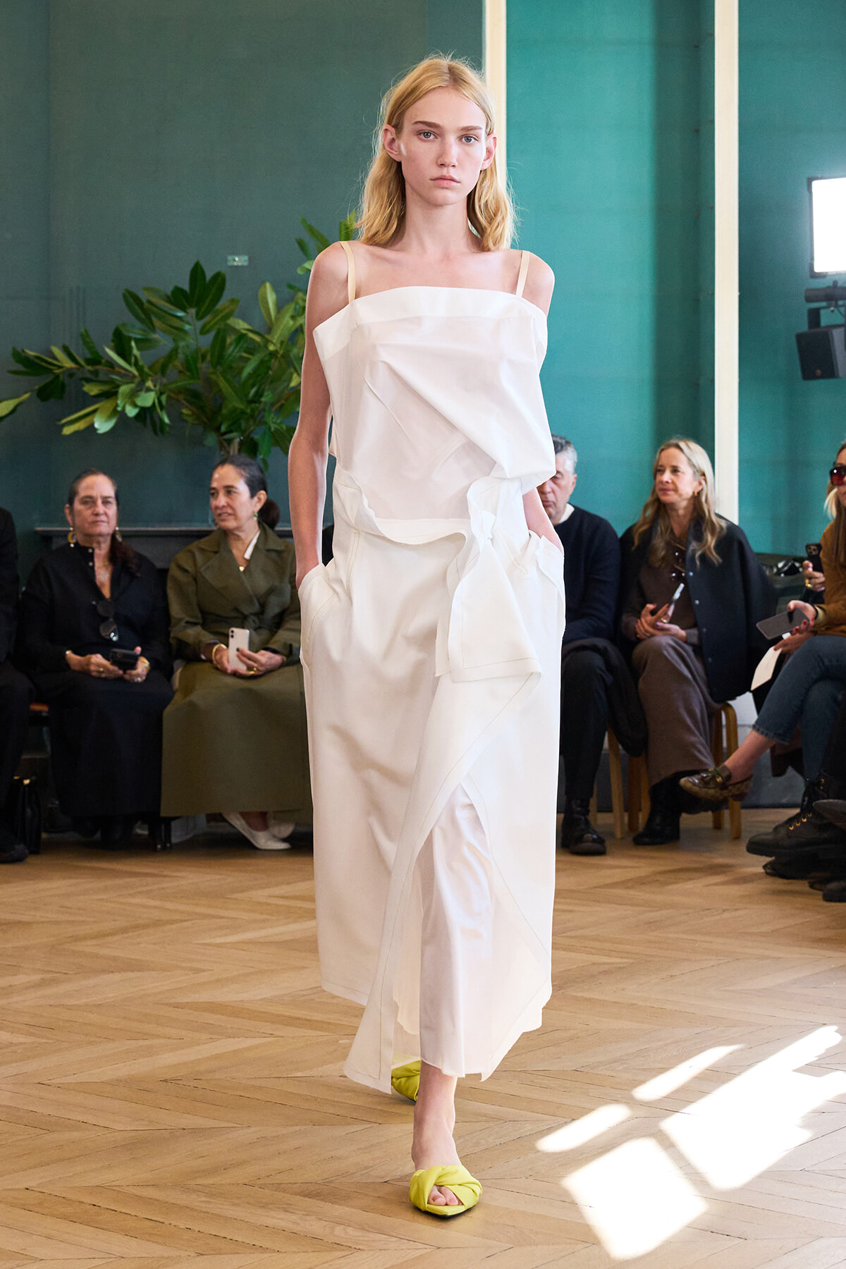 Model walks a fashion runway in a white draped dress, neon-yellow sandals, and a neutral expression, with seated audience in the background.