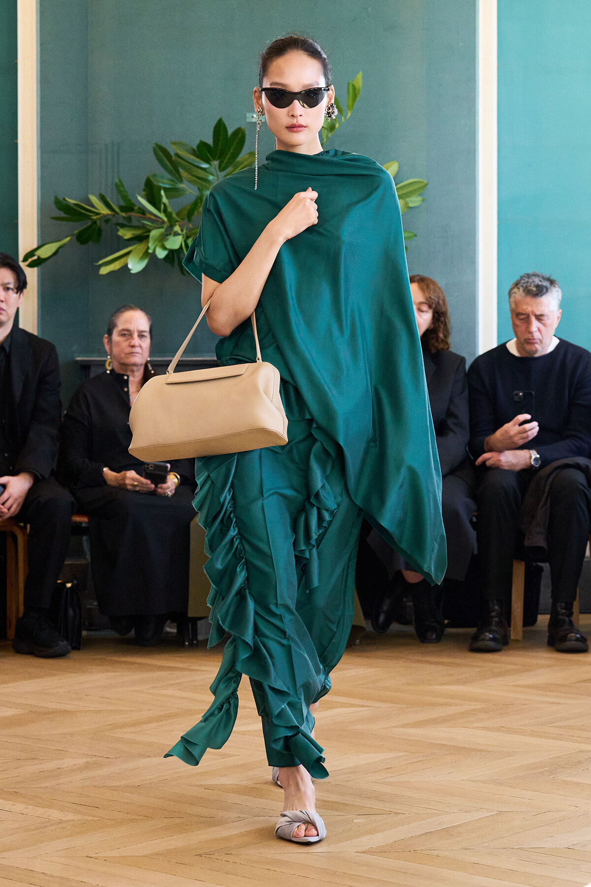 Model walks a runway in an emerald green, draped dress with a ruffled train, beige handbag, and dark sunglasses.