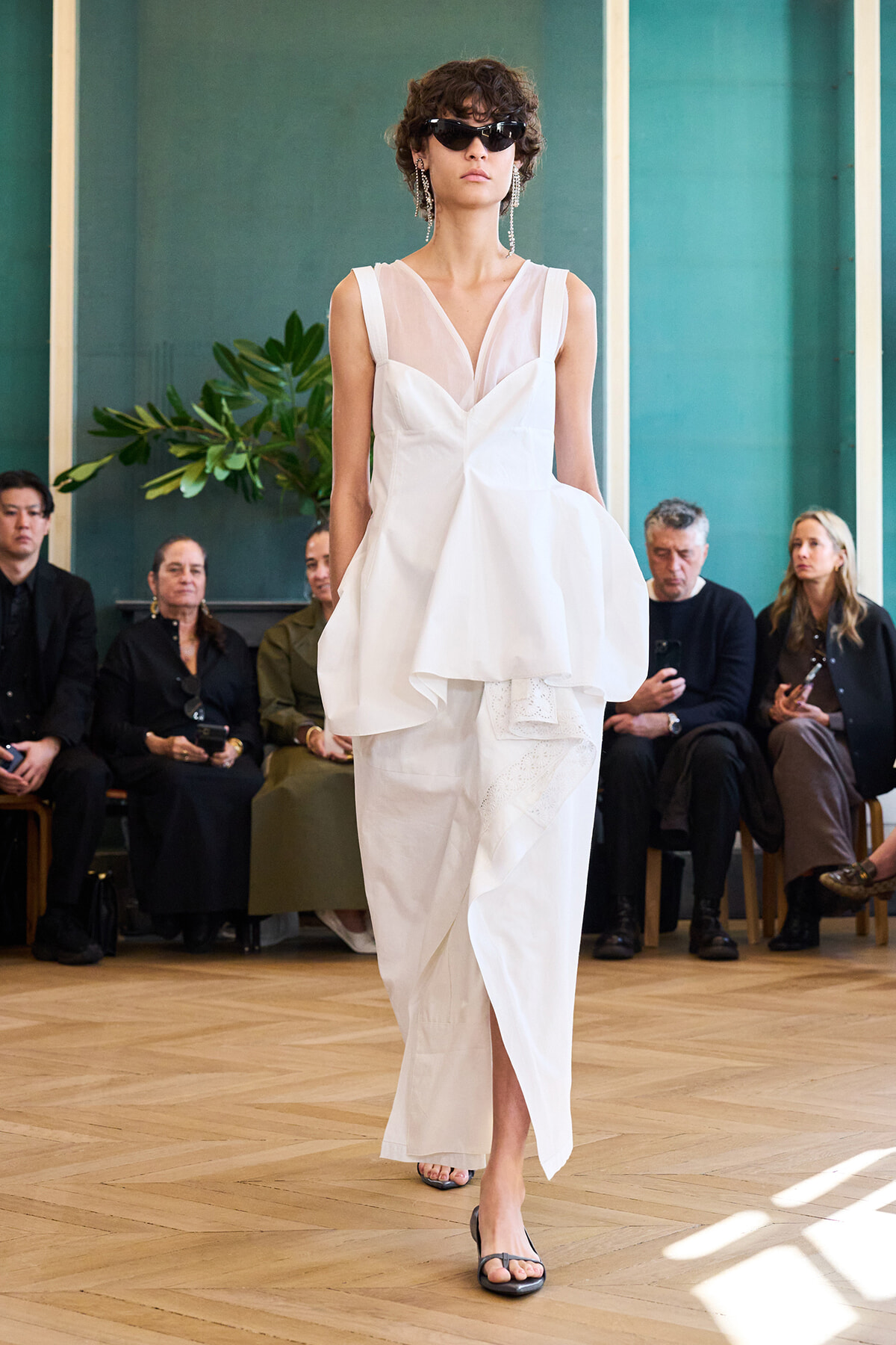 Model walks the runway in a white layered dress with a sheer bodice, black sunglasses, and dangling earrings.