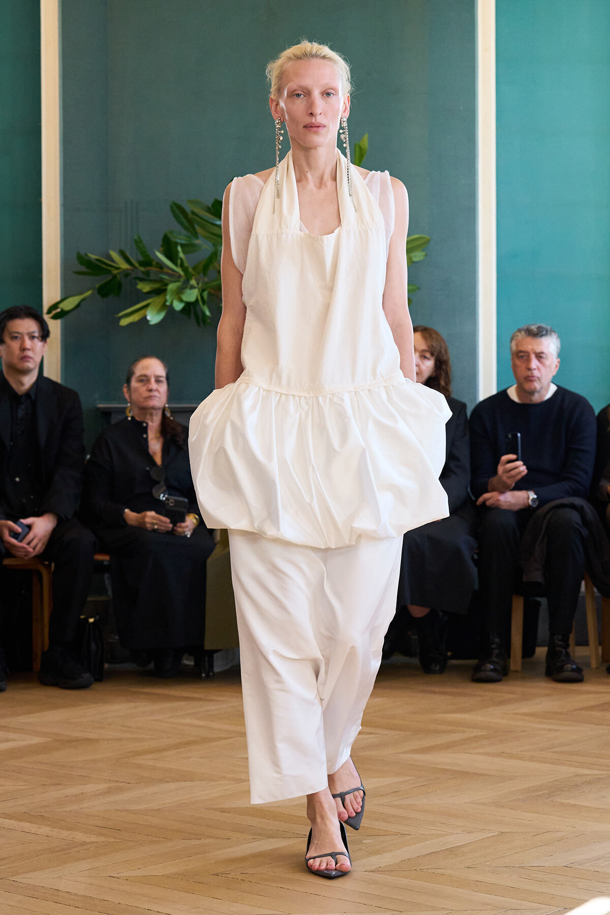 Model walks a white, sculptural outfit with a voluminous peplum over slim pants down a runway, audience seated in a gallery behind.