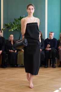 Model walking a runway in a two-tone sleeveless top and black pencil skirt, carrying a black shoulder bag; audience watches in background.