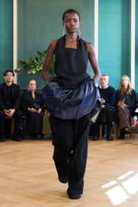Model walks a runway in a black top and oversized dark blue draped bottom; dramatic earrings, as seated audience observes in a gallery-like room.