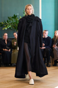 Model walks a fashion show wearing a long black layered cape over black outfit with white slides; audience seated in background.
